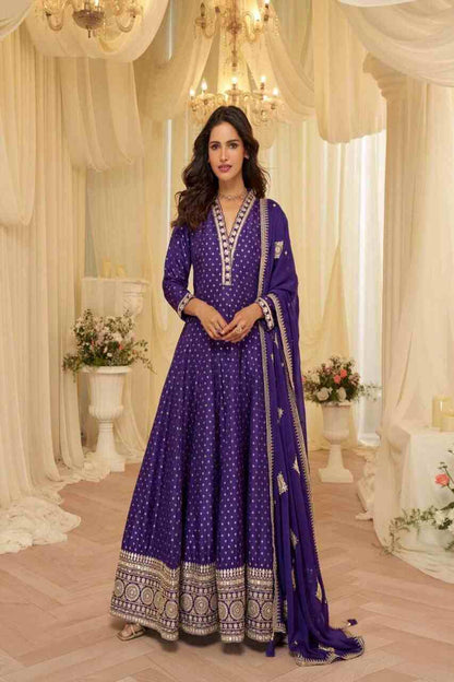Woman in a purple traditional outfit standing in a decorated room with chandeliers and floral arrangements.